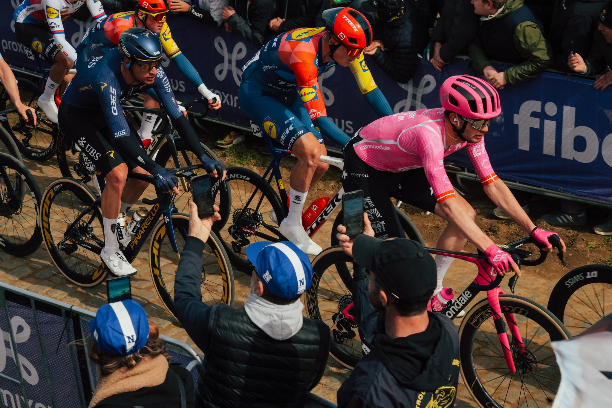 Three cyclists from INEOS Grenadiers, Lidl-Trek, and EF Education-EasyPost riding on a dusty gravel road, cheered by spectators.
