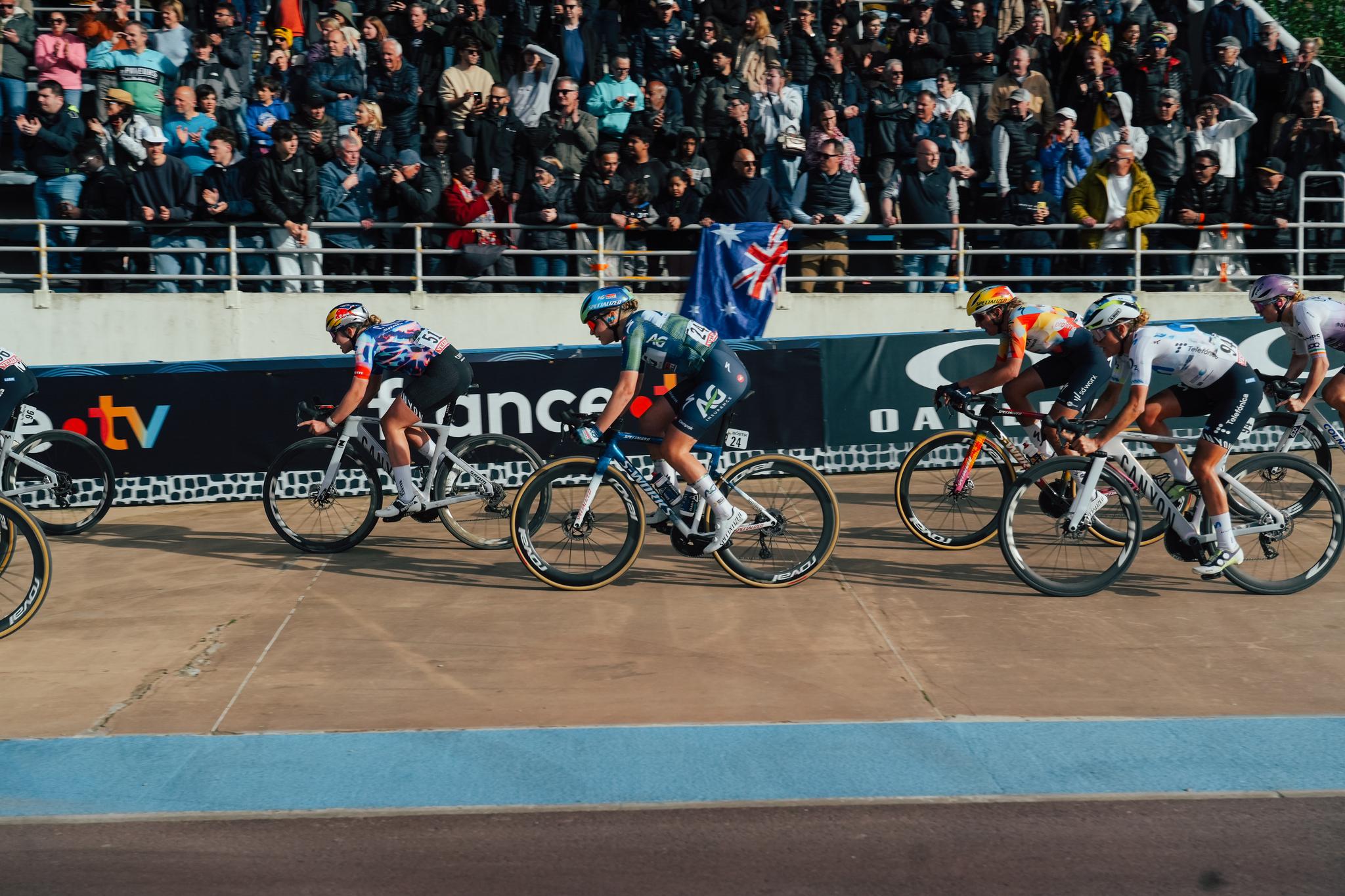 Female cyclists from AG Insurance – Soudal Team and EF Education-EasyPost race intensely on a velodrome track, with spectators and an Australian flag in the background.
