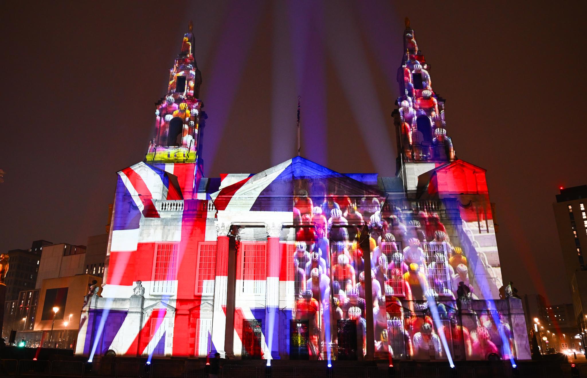 Night view of Leeds Civic Hall illuminated with a Union Jack flag and projected images of cyclists during the Tour de France 2027 Grand Depart route reveal.