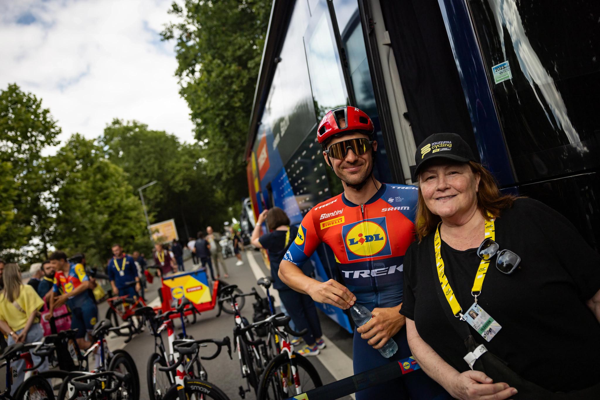 Julien Bernard of Lidl-Trek smiles alongside a Mummu Cycling guest beside the team bus at the Tour de France.